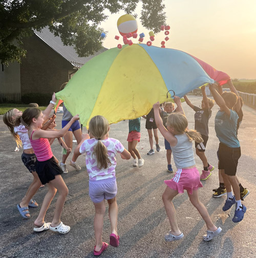 children work together  using a large, colorful tarp to toss several light-weight toys into the air